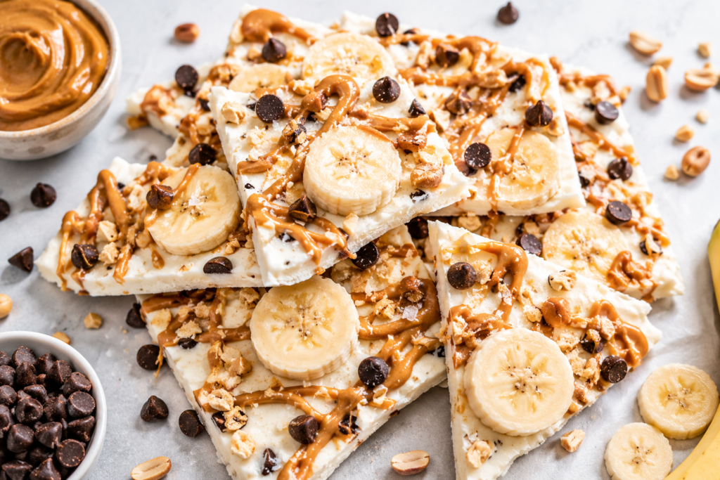 Peanut butter banana frozen yogurt bark with chocolate chips, banana slices, and chopped peanuts on a marble surface.